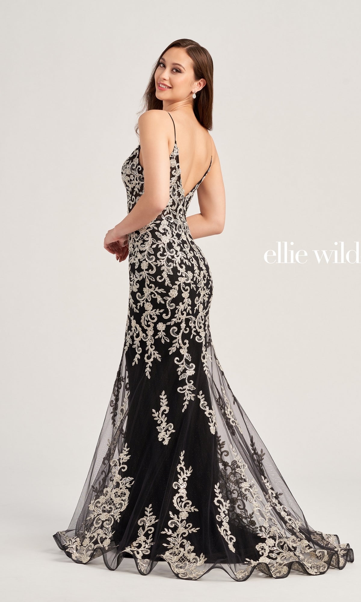 A woman poses in the Ellie Wilde Long Glitter-Lace Prom Dress EW35071, a fitted, floor-length black gown featuring silver embroidery and a low back, against a plain light background.